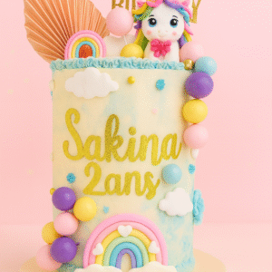 Gâteau d’anniversaire licorne arc-en-ciel avec nuages, boules colorées, éventail doré et prénom en lettres pailletées.