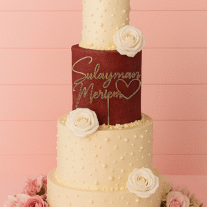 Gâteau de mariage à quatre étages ivoire perlé avec étage bordeaux, roses blanches et prénoms en or devant.