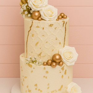 Gâteau à deux étages ivoire et or avec frise orientale en relief, texture dentelle, roses blanches, éventail doré, boules métalliques et feuille d’or.